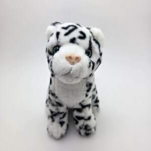 Può includere: Un peluche di leopardo delle nevi con corpo bianco e macchie nere. L'animale di peluche ha occhi verdi e naso rosa. È seduto, progettato per assomigliare a un vero leopardo delle nevi.