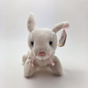 以下が含まれることがあります： ピンクのひげとピンクの鼻を持つ白いウサギのぬいぐるみ。ぬいぐるみは黒い目を持ち、耳にタグが付いています。ウサギは白い表面に横たわっています。
