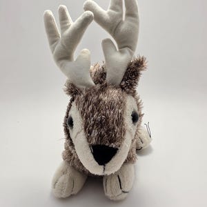 May include: A plush reindeer toy featuring a textured brown and white faux fur body, large cream-colored fabric antlers, black plastic eyes, and a black nose. The stuffed animal is in a lying position with its front paws extended forward.