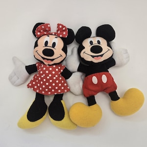 May include: A plush toy set featuring Mickey Mouse and Minnie Mouse. Mickey is wearing red shorts with white polka dots and yellow shoes. Minnie is wearing a red dress with white polka dots and yellow shoes. Both have black ears and black eyes.
