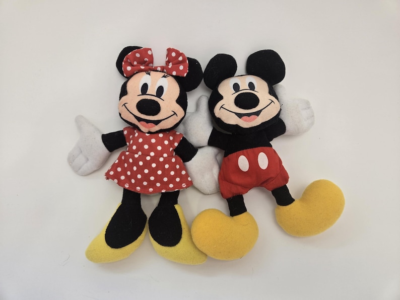 May include: A plush toy set featuring Mickey Mouse and Minnie Mouse. Mickey is wearing red shorts with white polka dots and yellow shoes. Minnie is wearing a red dress with white polka dots and yellow shoes. Both have black ears and black eyes.