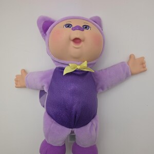 May include: A plush doll in a purple cat costume with a yellow bow tie. The doll has a round face, blue eyes, and is smiling with its arms outstretched. The costume has a sparkly purple torso and a tail.