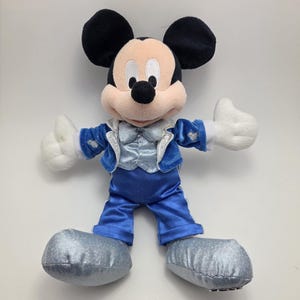 May include: A plush Mickey Mouse toy wearing a blue satin suit with a silver vest and matching shoes. The toy has black ears, a tan face, and white gloves. The toy is standing with its arms outstretched.