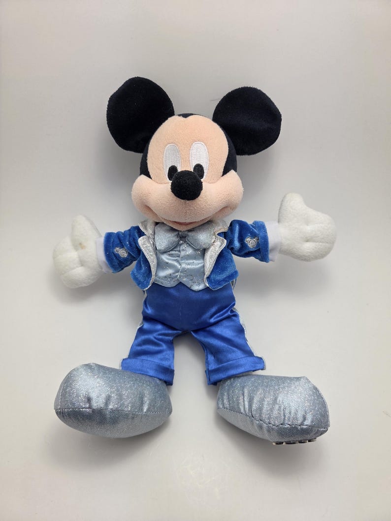 May include: A plush Mickey Mouse toy wearing a blue satin suit with a silver vest and matching shoes. The toy has black ears, a tan face, and white gloves. The toy is standing with its arms outstretched.