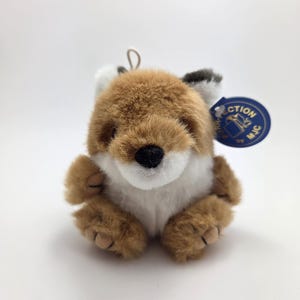 May include: A plush fox toy with brown, white, and black fur. The fox has a black nose and a blue tag attached. The toy is sitting upright with its paws in front. The fox is a cute and cuddly stuffed animal.