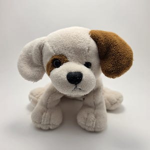 May include: A soft toy dog in cream with brown patches around one eye and on one ear. It has a black nose and eyes. The toy dog is positioned on a white surface.