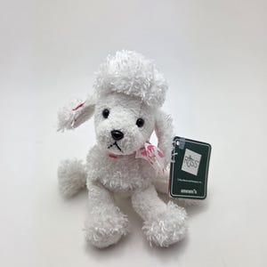 May include: A white plush poodle toy with black eyes and a black nose. The poodle has a pink ribbon around its neck and a tag that reads "ROSS". The toy is sitting on a white surface.