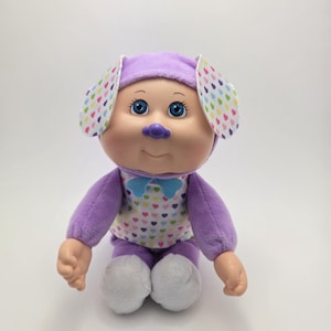 Puede incluir: Muñeca de peluche con forma de perro, con gorro y cuerpo morados, orejas blancas y una camisa adornada con corazones de colores. La muñeca tiene ojos azules, nariz morada y pajarita azul.