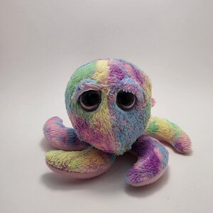 May include: A plush octopus toy featuring a tie-dye pattern with blended shades of pink, blue, yellow, and green. The stuffed animal has a soft, fuzzy texture, large dark brown eyes, and multiple curved tentacles extending from its round head.