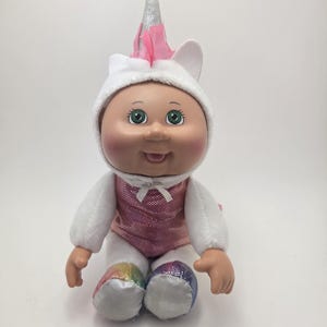 May include: A Cabbage Patch Kid doll dressed as a unicorn. The doll wears a white plush outfit with a pink sequined vest and a unicorn horn. The doll's feet are rainbow-colored, and it has green eyes.