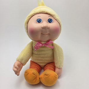 Peut inclure: Une poupée Cabbage Patch Kids vintage vêtue d'un costume de poussin jaune. La poupée a les yeux bleus, les joues roses et un nœud rose à pois blancs. La tenue de la poupée comprend un pantalon rayé orange et jaune et des pieds orange.