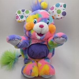 Könnte beinhalten: Ein farbenfrohes Plüschtier mit einem Batikmuster in Rosa, Gelb, Lila und Blau. Das Spielzeug hat große Ohren mit weißem Hintergrund und bunten Spiralmustern. Es hat einen lila Bauch, eine rosa Nase und einen grün-gelben Schwanz.