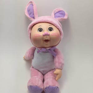 May include: A vintage Cabbage Patch Kids doll wearing a pink bunny costume with lavender accents. The doll has rosy cheeks, a pink nose, and a lavender bow. The bunny ears are lined with lavender fabric.