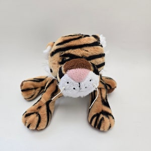 Puede incluir: Un juguete de peluche de tigre con rayas marrones y negras, una nariz rosa y bigotes blancos. El juguete est&aacute; acostado boca abajo con las patas extendidas.