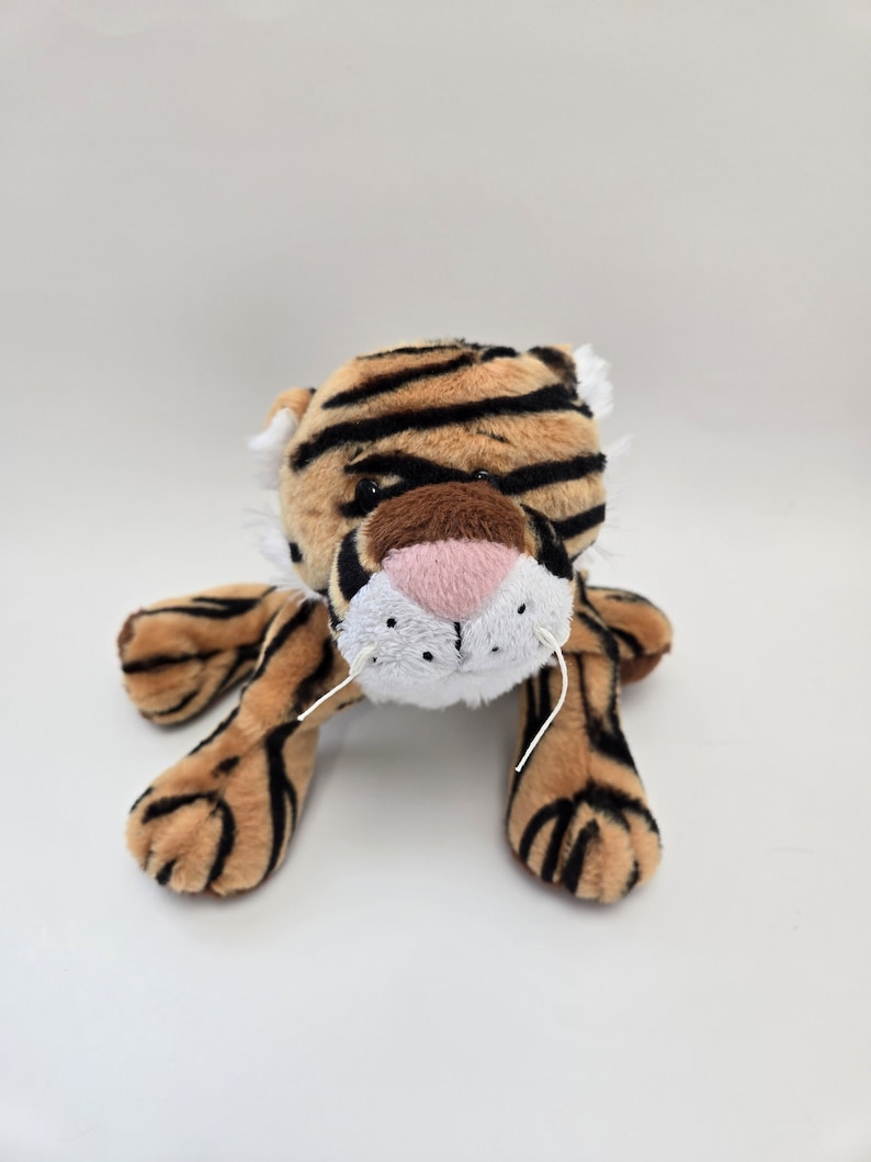Puede incluir: Un juguete de peluche de tigre con rayas marrones y negras, una nariz rosa y bigotes blancos. El juguete est&aacute; acostado boca abajo con las patas extendidas.
