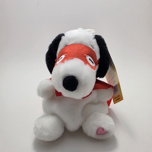 May include: A plush white dog toy with black ears, a black nose, and a red mask and cape. The dog has a pink heart on one paw. The toy is sitting upright.