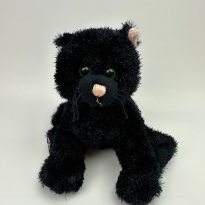 Peut inclure: Une peluche noire en forme de chat avec des yeux verts et un nez rose. Le chat est assis droit, les pattes avant tendues. Le jouet a de longues moustaches noires et une texture duveteuse.