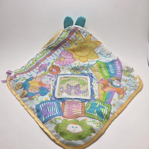 May include: A Cabbage Patch Kids baby blanket with a yellow satin border. The blanket features colorful illustrations of Cabbage Patch Kids characters, flowers, and blocks spelling out letters. The blanket has a small window with the text "Peek a Boo".