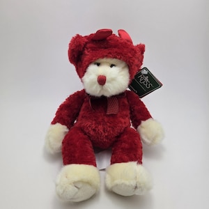 May include: A red teddy bear toy with a matching hat and scarf. The bear has a white face, paws, and feet. A black tag with the brand name "Russ" is attached to the bear.