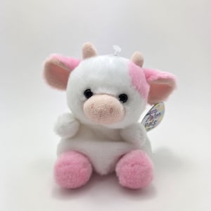 May include: A white and pink plush cow with black eyes and a pink nose. The cow is sitting with its legs crossed and has a tag that says "Palmo Pals".