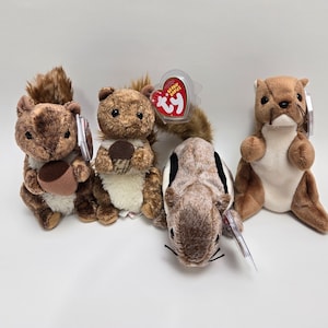 May include: Four plush squirrel toys in various poses. The squirrels are primarily brown with white and tan accents. Two squirrels hold acorns. Each toy has a tag attached. The toys are on a white surface.
