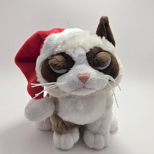 May include: A plush cat toy featuring white and brown fur with a grumpy facial expression, wearing a soft red Santa hat. The cat has dark brown patches around its eyes, a small pink nose, and long white whiskers extending from its muzzle.