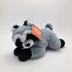May include: A plush grey raccoon with black eyes and a black nose. The raccoon is lying on its belly with its paws outstretched. The raccoon has a tag attached to its back that says "Wild Republic".