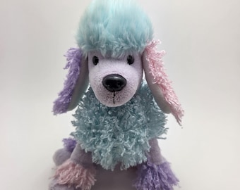 Op de afbeelding: Een pluche poedel speelgoed in pastelkleuren. De poedel heeft een lichtblauwe kop, een bijpassende fluffy kraag en een lavendelkleurig lichaam met roze en paarse accenten op de oren en poten. Het speelgoed heeft een zwarte neus.