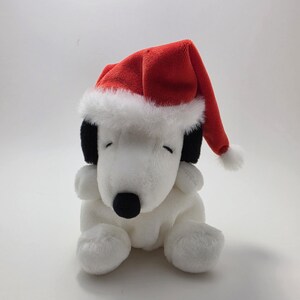 May include: A plush white dog toy with black ears and a black nose, wearing a red Santa hat with white trim and a small white pom-pom. The toy is sitting on a white surface.