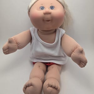 Peut inclure: Une poupée Cabbage Patch Kid vintage avec des cheveux blonds et des yeux bleus. La poupée porte un débardeur blanc et un short rouge. La poupée est assise, les bras tendus.