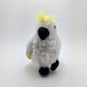 Può includere: Un giocattolo di cacatua bianco di peluche con cresta gialla, becco nero e piedi neri. L'animale di peluche ha una consistenza soffice ed è posto su uno sfondo bianco. Questo giocattolo è adatto ai bambini.