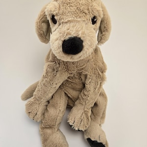 May include: A plush tan-colored dog toy with black eyes and a black nose. The dog is sitting with its paws tucked in.