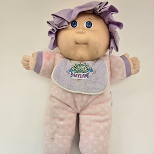 May include: A vintage Cabbage Patch Kids doll with a soft, plush body. The doll has a light skin tone, blue eyes, and a lavender bonnet. It wears a pink outfit with white star patterns and a bib that says "Babyland".