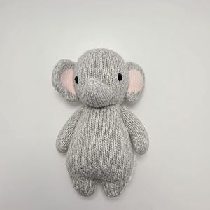 May include: A small, hand-knitted elephant toy made from light gray yarn. The plush features a rounded head, two large ears with soft pink inner accents, a trunk, and two small black embroidered eyes. The body is plump with short arms and legs.