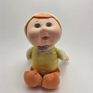 May include: A vintage Cabbage Patch Kids doll dressed in a yellow and orange outfit. The doll has a soft, plush body, green eyes, and a smiling expression. It is wearing a yellow bonnet with an orange brim and orange feet.