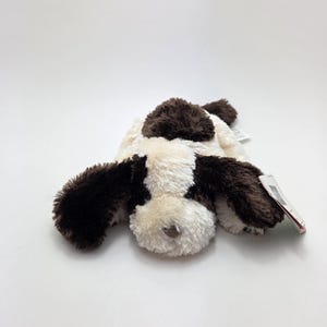 May include: A plush dog toy in a lying position, featuring a brown and white spotted pattern. The toy has floppy ears and a soft, fluffy texture. A small tag is attached to the toy.