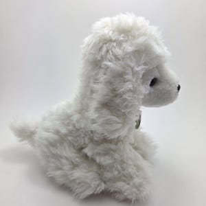 May include: A white plush poodle toy with a fluffy, textured coat. The toy has black eyes and a small, round nose. The poodle is sitting, with its tail curled slightly. A small tag is visible on the collar.