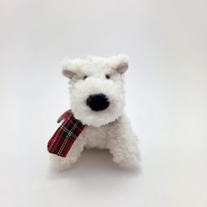 May include: White plush toy dog wearing a red and black plaid scarf. The dog is sitting with its head tilted slightly to the side.