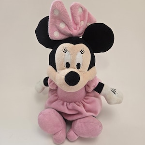 Puede incluir: Juguete de peluche rosa Minnie Mouse con orejas negras, un lazo rosa con lunares blancos y un vestido rosa.