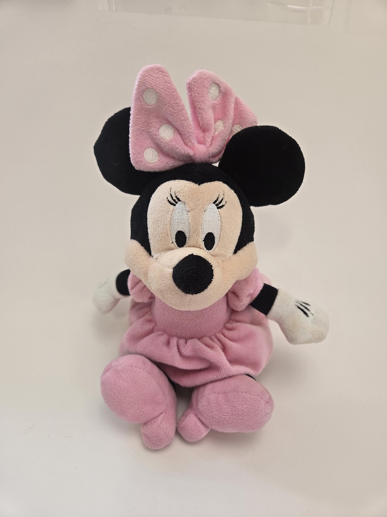 Puede incluir: Juguete de peluche rosa Minnie Mouse con orejas negras, un lazo rosa con lunares blancos y un vestido rosa.
