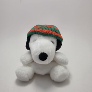 May include: A plush white Snoopy toy wearing a green and orange striped knit cap. The toy has black ears and a black nose. The Snoopy plush is sitting down with its arms and legs extended.