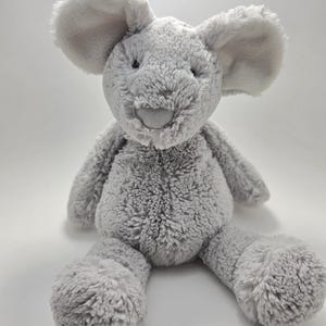 May include: A plush stuffed animal in the shape of a mouse with soft, light gray textured fur. The mouse features large, rounded ears, a small smooth gray nose, and dark embroidered eyes. It is depicted in a seated position with its arms and legs extended forward.