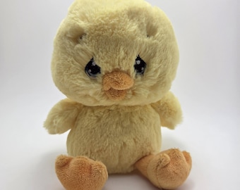 May include: A plush yellow chick with a tuft of yellow feathers on its head. The chick has a light orange beak and feet, and black eyes. The chick is sitting on a white surface.