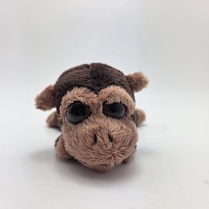 Peut inclure: Une peluche singe avec une fourrure brun foncé sur le dessus de la tête et une fourrure beige plus claire sur le visage et les oreilles. L'animal en peluche a de grands yeux noirs brillants et ronds ainsi qu'un petit museau texturé.