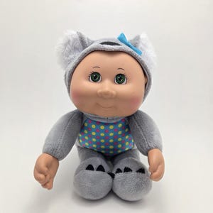 May include: A gray plush koala baby doll with a blue bow and a yellow, pink, and blue polka dot pattern on its chest. The doll has green eyes and a smiling face.
