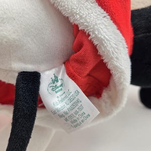 May include: A plush toy of Mickey Mouse dressed as Santa Claus. The toy is wearing a red and white Santa suit with a white fur trim. The tag on the toy reads "Disney Baby" and includes information about the manufacturer and country of origin.
