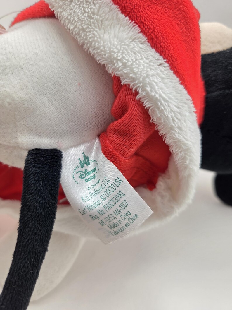 May include: A plush toy of Mickey Mouse dressed as Santa Claus. The toy is wearing a red and white Santa suit with a white fur trim. The tag on the toy reads "Disney Baby" and includes information about the manufacturer and country of origin.