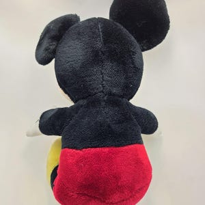 May include: A plush Mickey Mouse toy, viewed from the back. The toy features a black head and ears, a black body, red shorts, and yellow feet. The toy is made of soft, plush material.