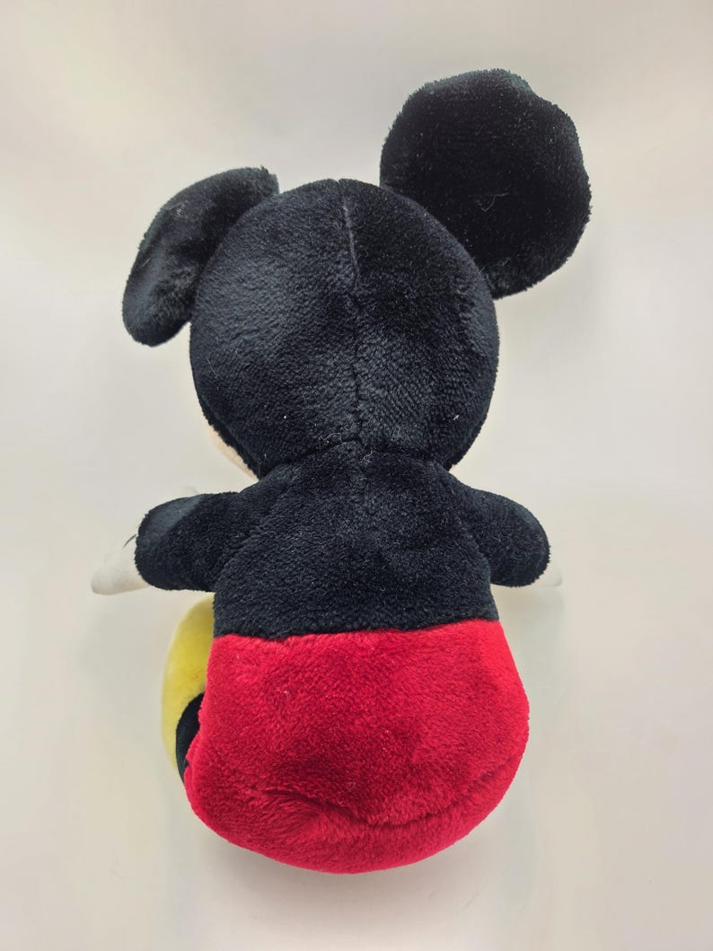 May include: A plush Mickey Mouse toy, viewed from the back. The toy features a black head and ears, a black body, red shorts, and yellow feet. The toy is made of soft, plush material.