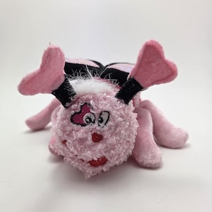 May include: A plush pink bug toy with heart-shaped antennae and six legs. The bug has a curly texture on its head and a black and pink patterned back. The toy has a smiling red mouth and a heart-shaped design on its face.